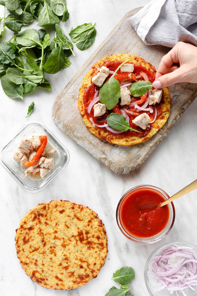 Simple Cauliflower Pizza Crust on a wooden cutting board. The cauliflower curst is surrounded by pizza ingredients on a white marble countertop.