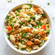 Chicken fried cauliflower rice served in a white bowl with tender chicken pieces, peas, carrots, herbs, and fluffy cauliflower rice on a light marble background.