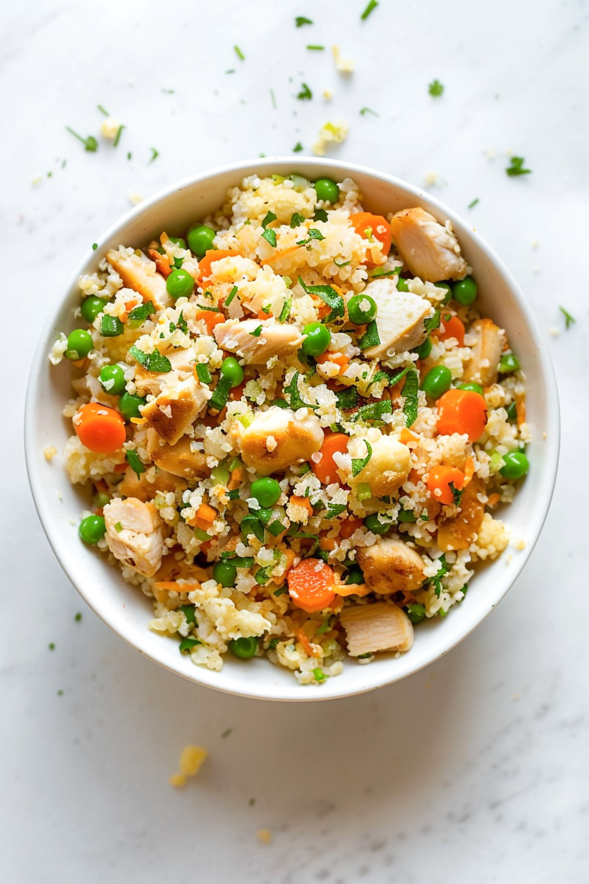 Chicken fried cauliflower rice served in a white bowl with tender chicken pieces, peas, carrots, herbs, and fluffy cauliflower rice on a light marble background.