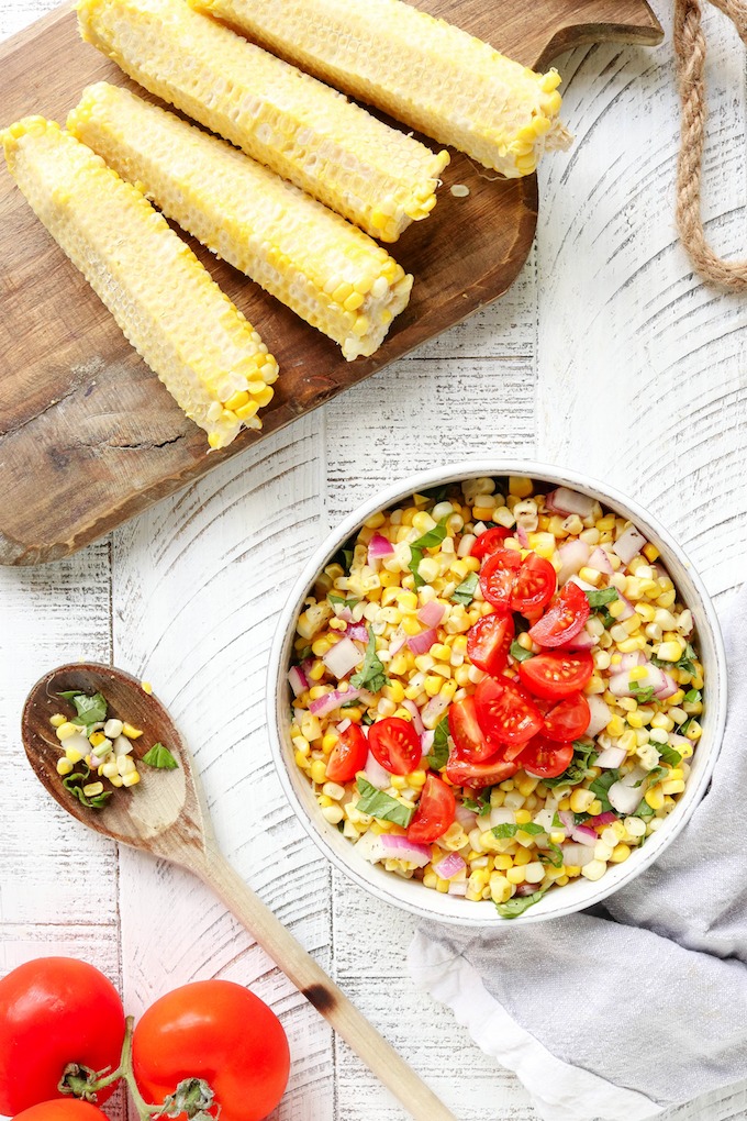 10-Minute Fresh Corn Salad in a white bowl. Corn cobs and a wooden spoon are arranged on a white wooden background.