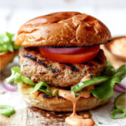 Juicy Asian turkey burger on a toasted brioche bun with lettuce, tomato slices, red onion, and creamy sriracha mayo dripping down the side.