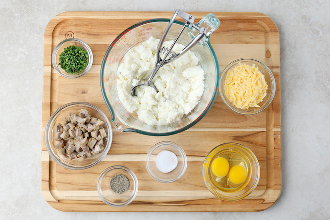 Mashed Potato Puff Ingredients on a wooden cutting board. Mashed potatoes, chives, sausage, eggs, salt, pepper and cheese.
