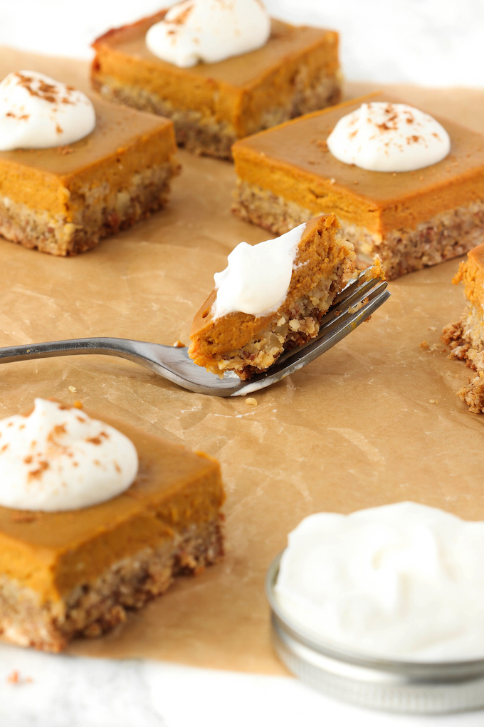 Healthy pumpkin pie bars scattered on a wooden cutting board. Center is a fork with a bite of pie topped with whipped cream.