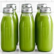Six glass bottles with silver metal caps, filled with green juice, lined up in a row on a white background.