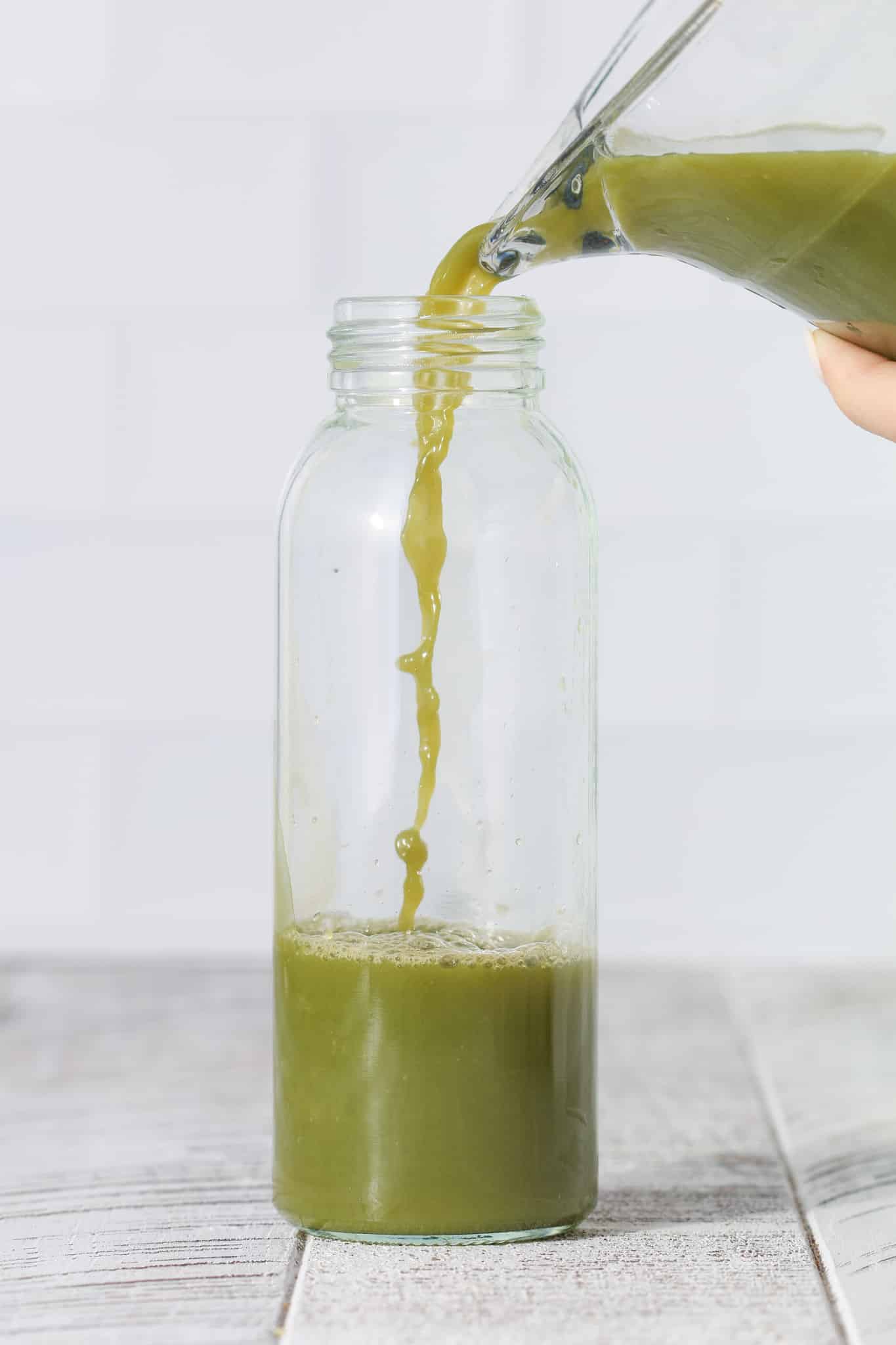 Green juice being poured from a bottle into a clear glass jar, with juice collecting at the bottom on a light-colored surface.