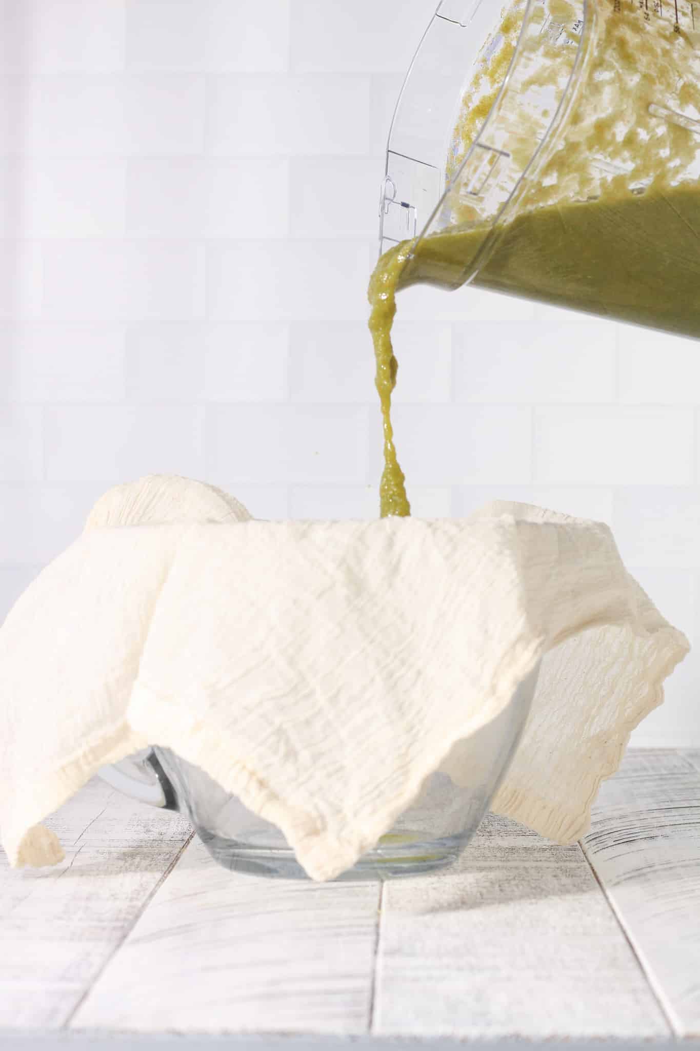 Green juice being poured onto a cloth-lined strainer set over a bowl, preparing juice for straining.