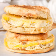 Close-up of two homemade sausage, egg, and cheese English muffin sandwiches stacked on parchment paper, with melted cheese and golden toasted muffins.