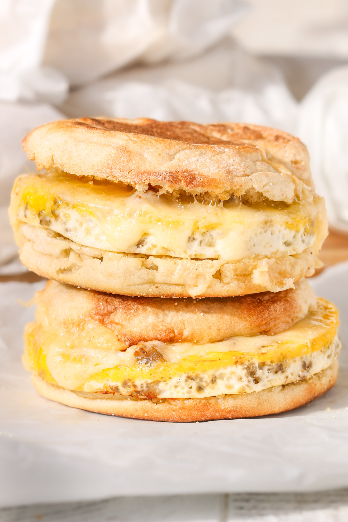 Close-up of two homemade sausage, egg, and cheese English muffin sandwiches stacked on parchment paper, with melted cheese and golden toasted muffins.