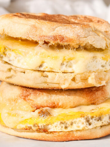 Close-up of two homemade sausage, egg, and cheese English muffin sandwiches stacked on parchment paper, with melted cheese and golden toasted muffins.