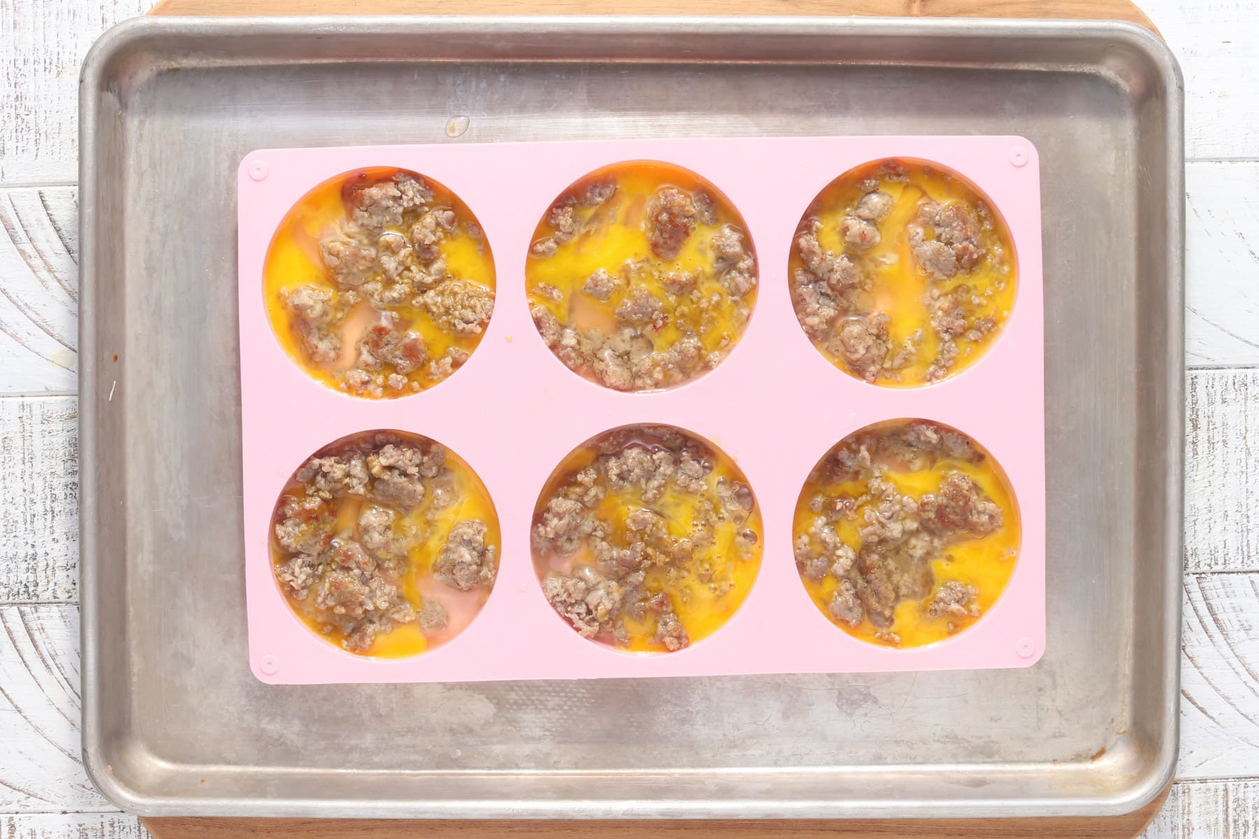 Overhead view of uncooked egg and sausage mixture in a pink silicone mold on a sheet pan, ready to bake for freezer breakfast sandwiches.