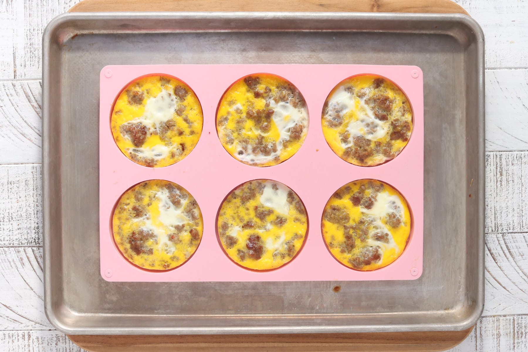 Overhead view of baked egg and sausage rounds in a pink silicone mold on a sheet pan, part of the prep process for freezer breakfast sandwiches.