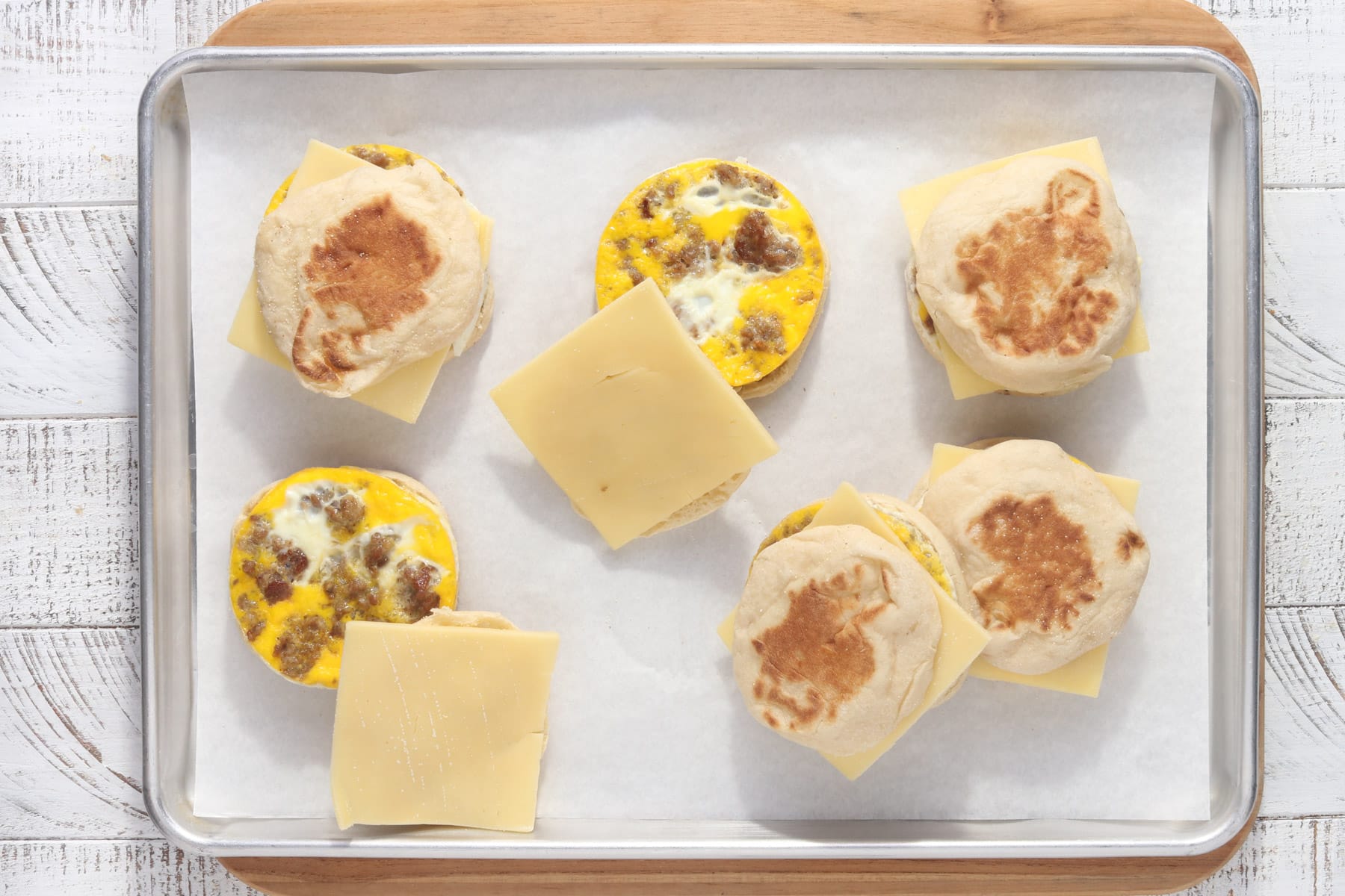 Overhead view of assembled freezer breakfast sandwiches on a sheet pan, featuring toasted English muffins, baked egg and sausage rounds, and slices of cheese ready to freeze.