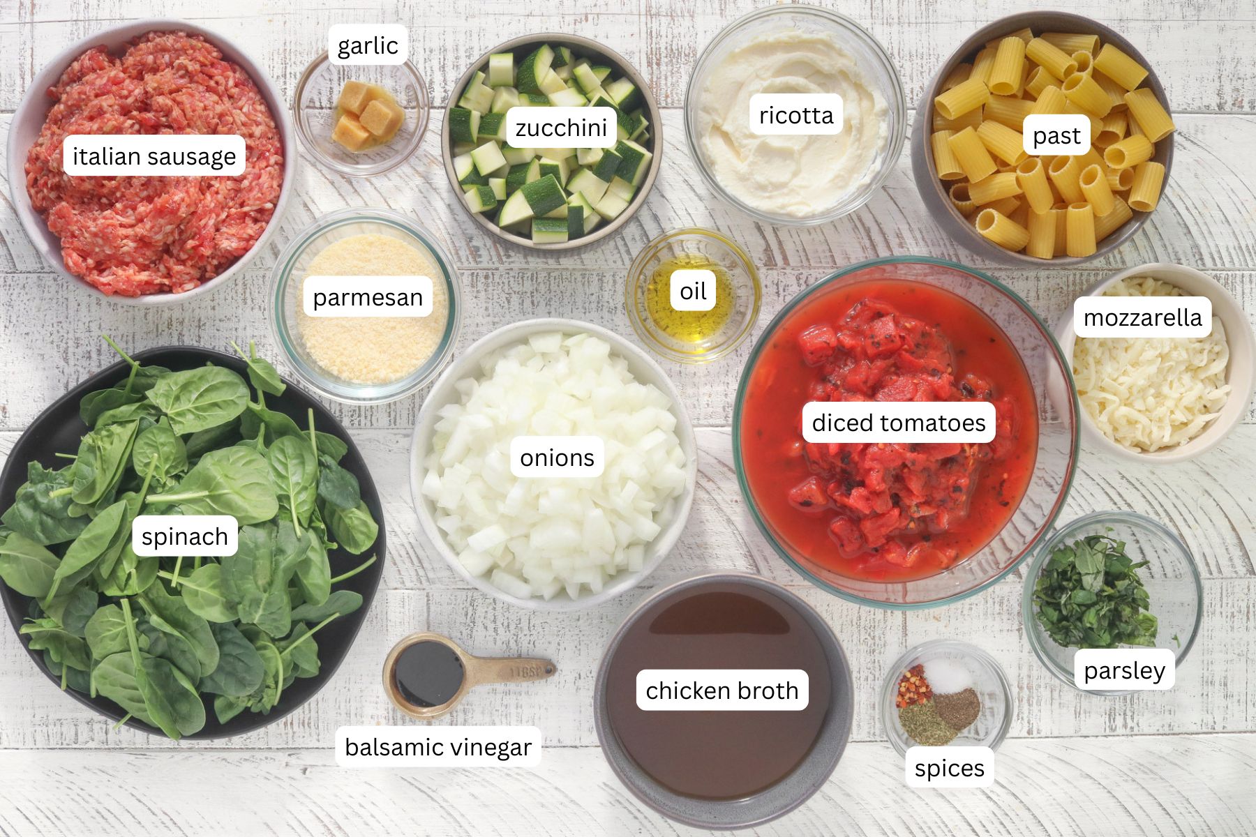Overhead flat lay of labeled ingredients for sausage and ricotta pasta arranged in glass and ceramic bowls on a white wood surface: Italian sausage, garlic, zucchini, ricotta, rigatoni pasta, parmesan, olive oil, diced tomatoes, mozzarella, spinach, onions, chicken broth, balsamic vinegar, spices, and parsley.