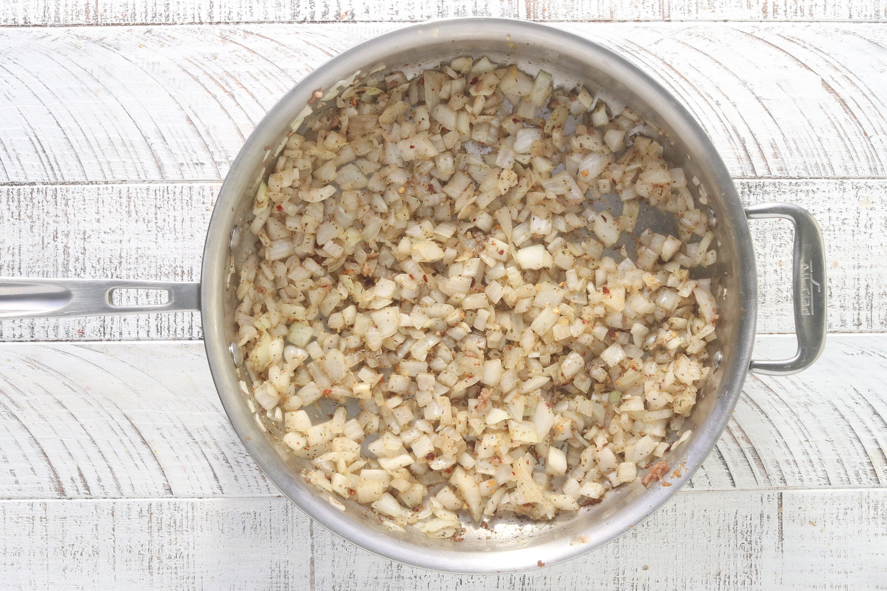Overhead view of diced onion, garlic, and spices cooking in a large stainless steel skillet on a white wood surface.