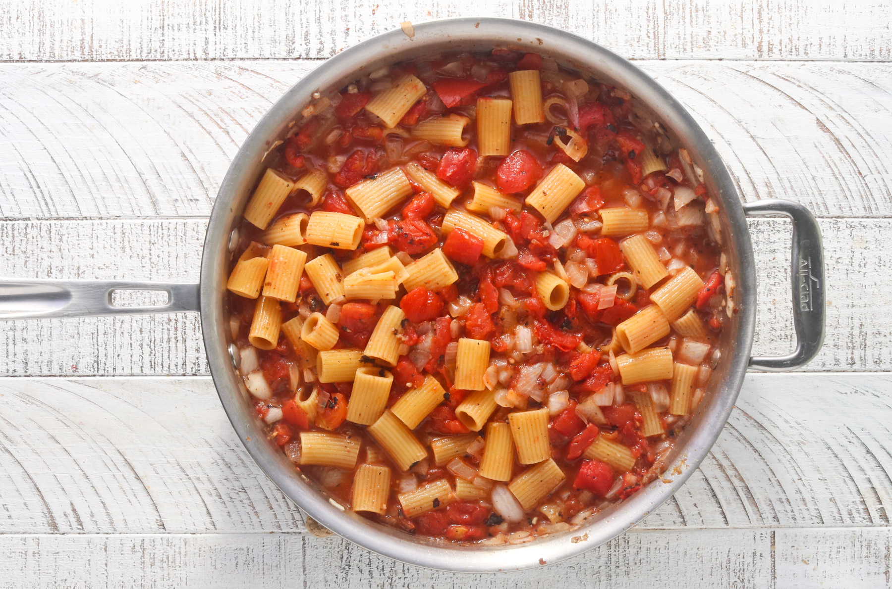 Overhead view of dry rigatoni, diced tomatoes, and chicken broth combined in a large stainless steel skillet before simmering, on a white wood surface.