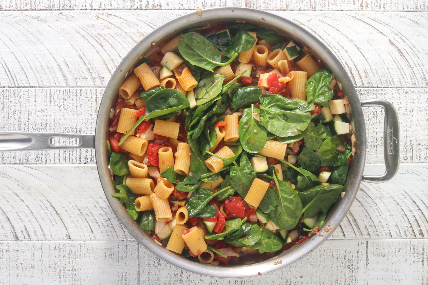 Overhead view of fresh spinach and chopped zucchini added to rigatoni and tomato sauce in a large stainless steel skillet, on a white wood surface.