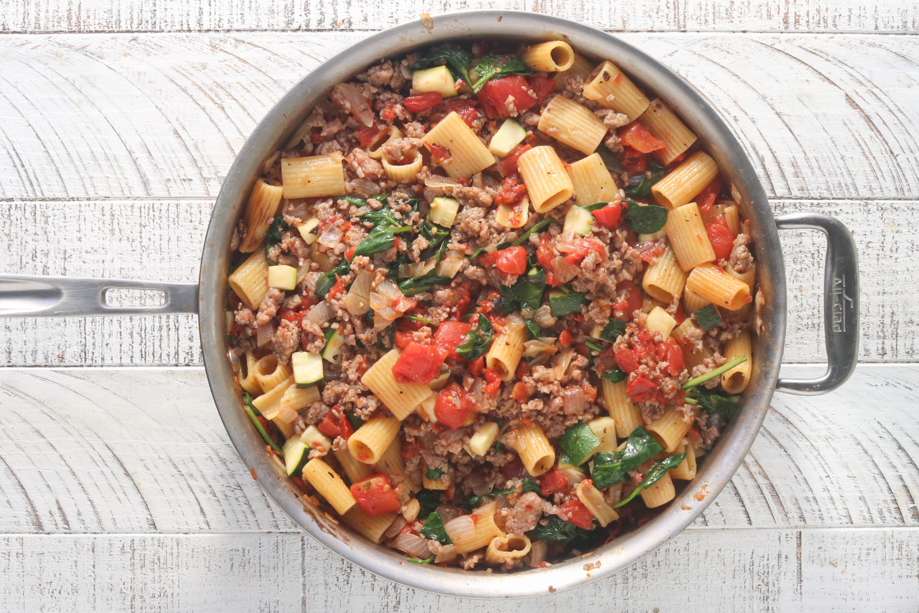 Overhead view of cooked Italian sausage stirred into rigatoni with tomatoes, zucchini, and wilted spinach in a large stainless steel skillet, on a white wood surface.