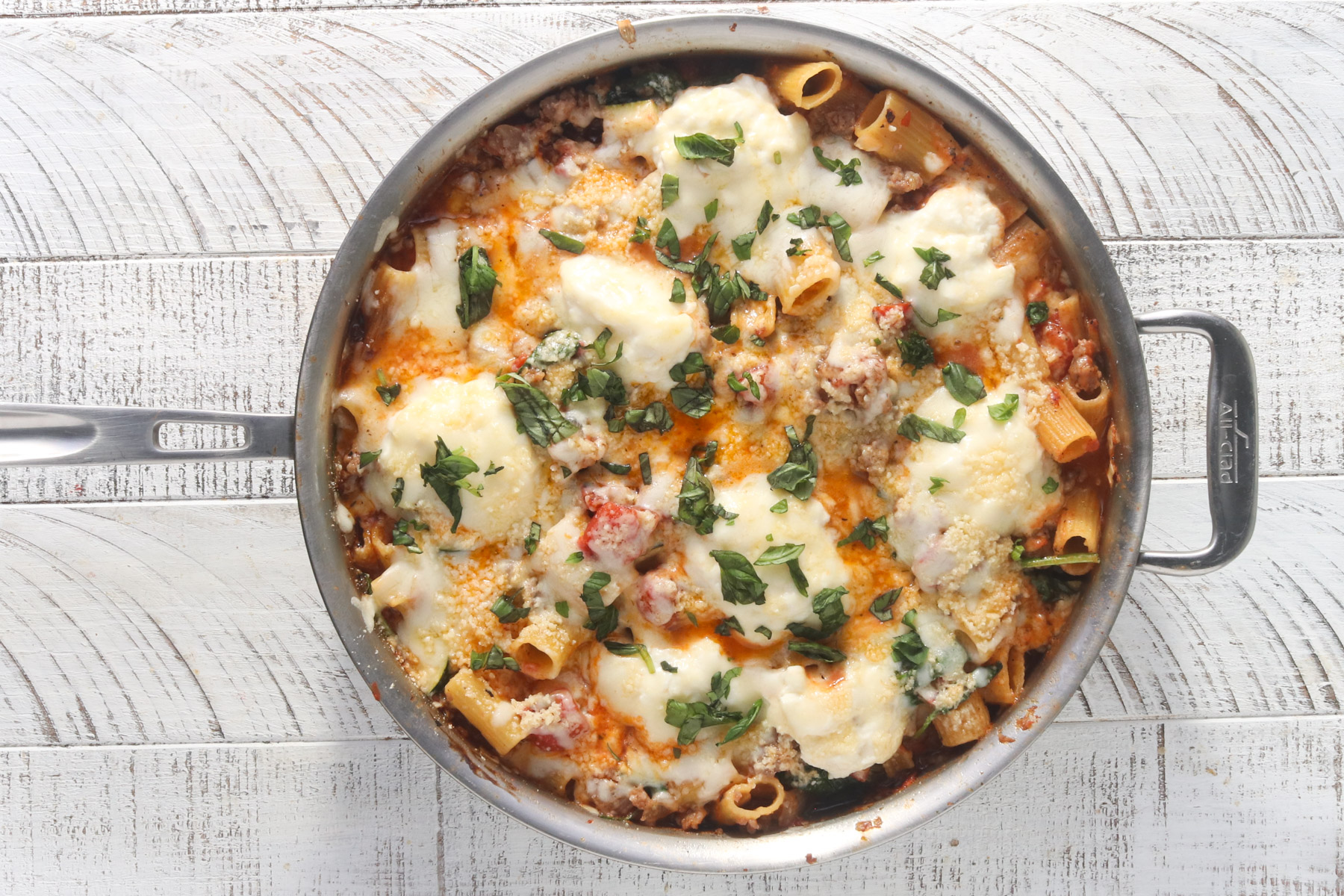 Overhead view of finished sausage and ricotta pasta in a large stainless steel skillet, topped with melted mozzarella, ricotta dollops, Parmesan, and fresh chopped basil, on a white wood surface.