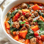 A hearty bowl of sweet potato sausage stew filled with chunks of tender orange sweet potatoes, crumbled browned sausage, wilted spinach, and diced tomatoes in a rich, flavorful broth. The stew is served in a white bowl on a rustic white wood surface with a linen napkin and a spoon in the background.