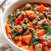 A hearty bowl of sweet potato sausage stew filled with chunks of tender orange sweet potatoes, crumbled browned sausage, wilted spinach, and diced tomatoes in a rich, flavorful broth. The stew is served in a white bowl on a rustic white wood surface with a linen napkin and a spoon in the background.