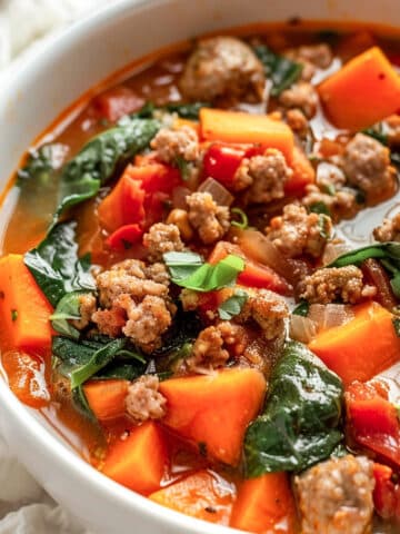 A hearty bowl of sweet potato sausage stew filled with chunks of tender orange sweet potatoes, crumbled browned sausage, wilted spinach, and diced tomatoes in a rich, flavorful broth. The stew is served in a white bowl on a rustic white wood surface with a linen napkin and a spoon in the background.