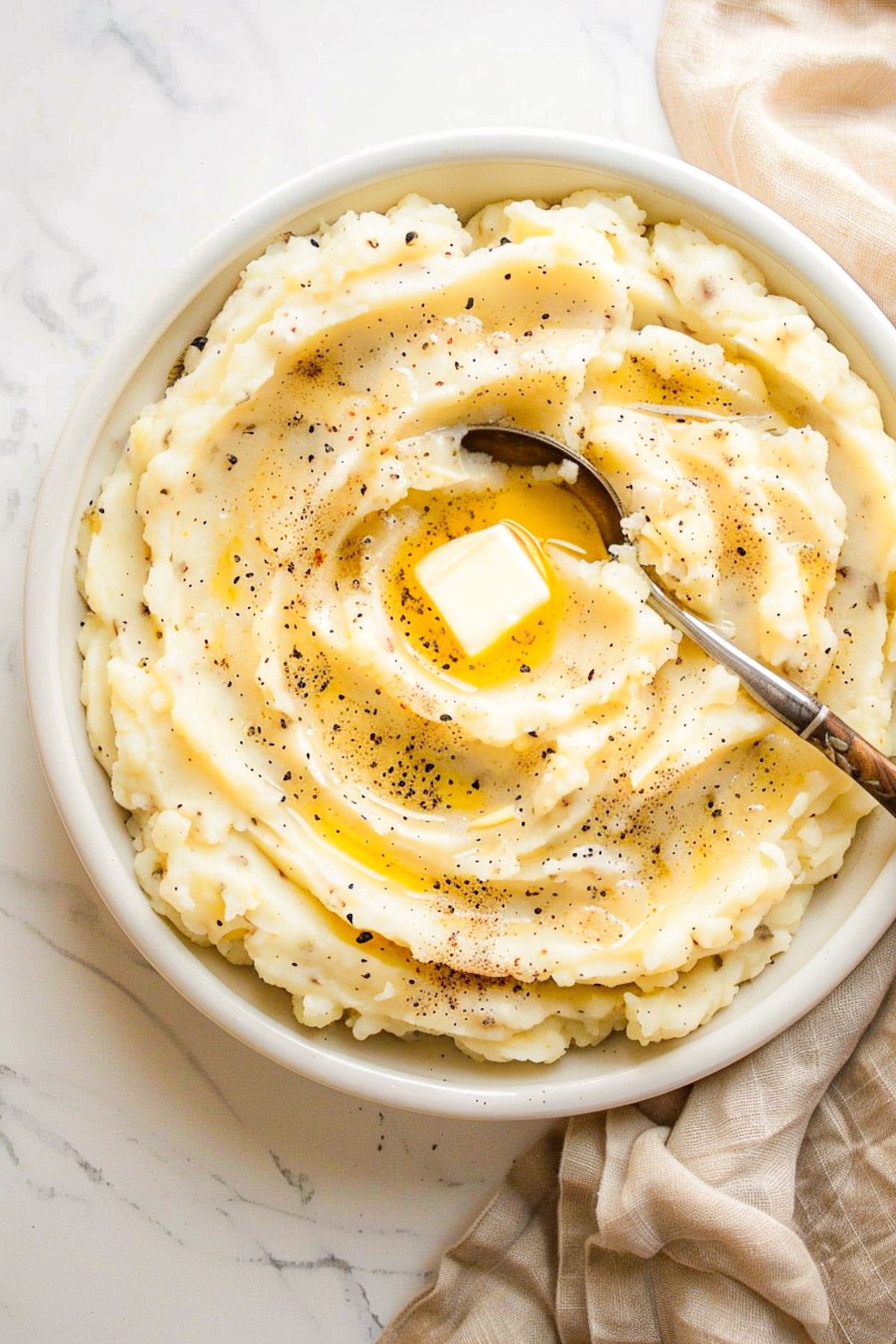 A bowl of creamy mashed potatoes topped with melted butter and cracked black pepper, with a spoon resting in the swirl, sitting on a light marble surface with a tan linen napkin.