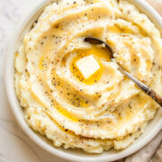 A bowl of creamy mashed potatoes topped with melted butter and cracked black pepper, with a spoon resting in the swirl, sitting on a light marble surface with a tan linen napkin.