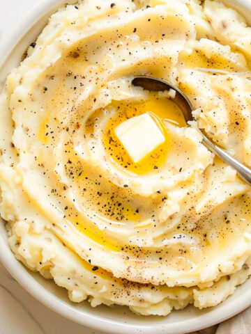 A bowl of creamy mashed potatoes topped with melted butter and cracked black pepper, with a spoon resting in the swirl, sitting on a light marble surface with a tan linen napkin.