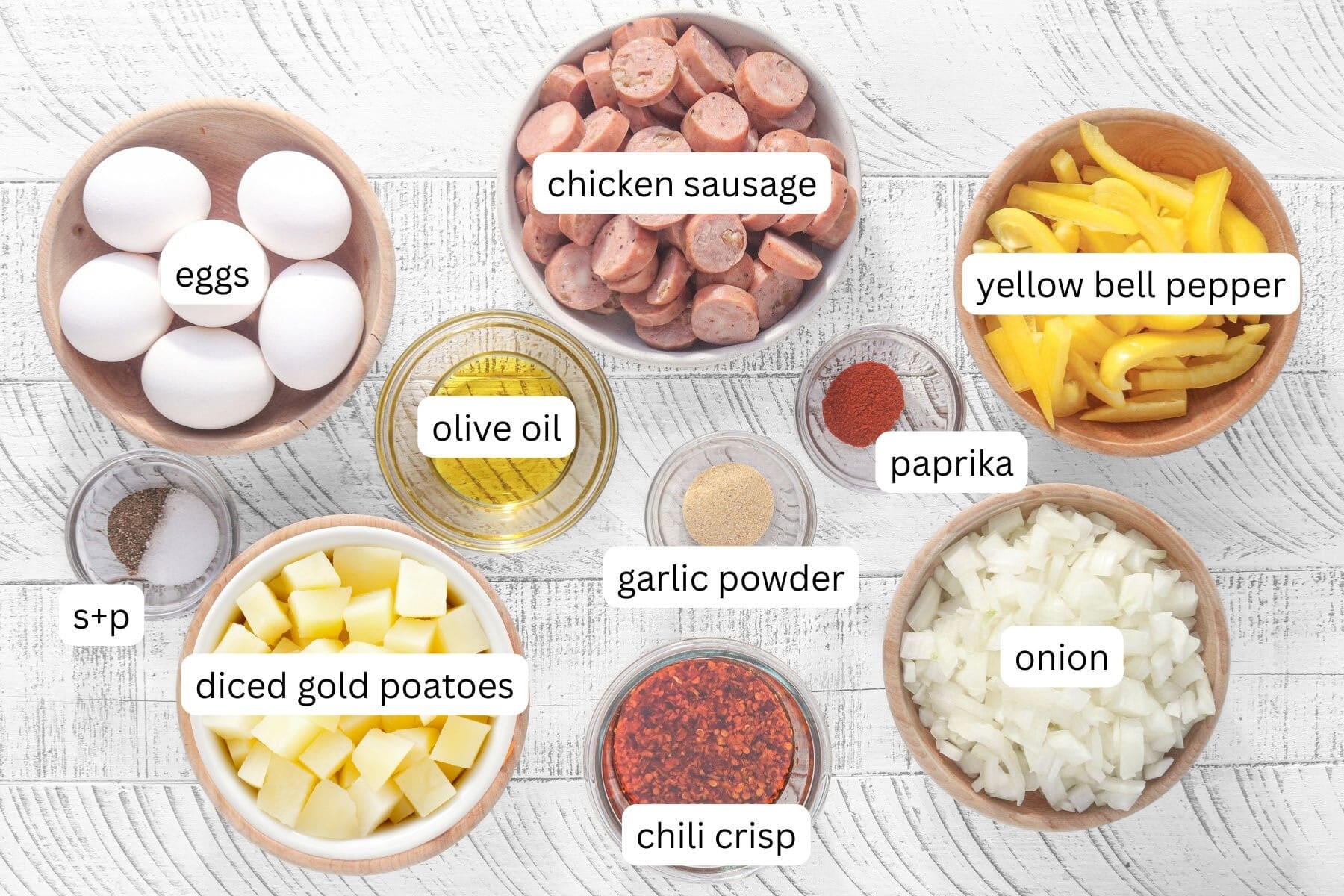 Overhead view of ingredients for eggs on a sheet pan with potatoes and sausage, including diced Yukon gold potatoes, chicken sausage, eggs, yellow bell pepper, onion, olive oil, garlic powder, paprika, salt and pepper, and chili crisp arranged in small bowls.