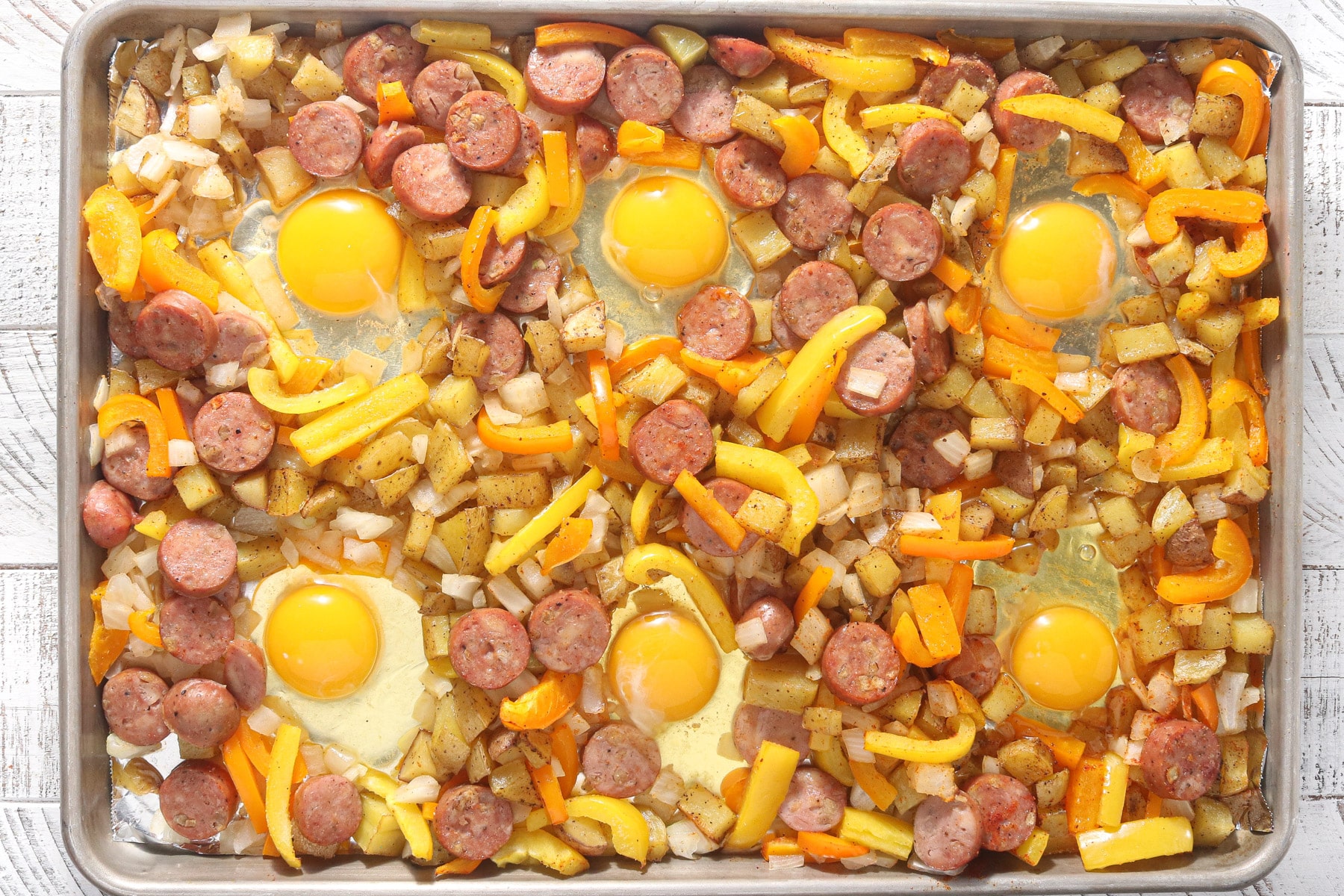 Eggs cracked into wells on a sheet pan with roasted potatoes, chicken sausage, and bell peppers.