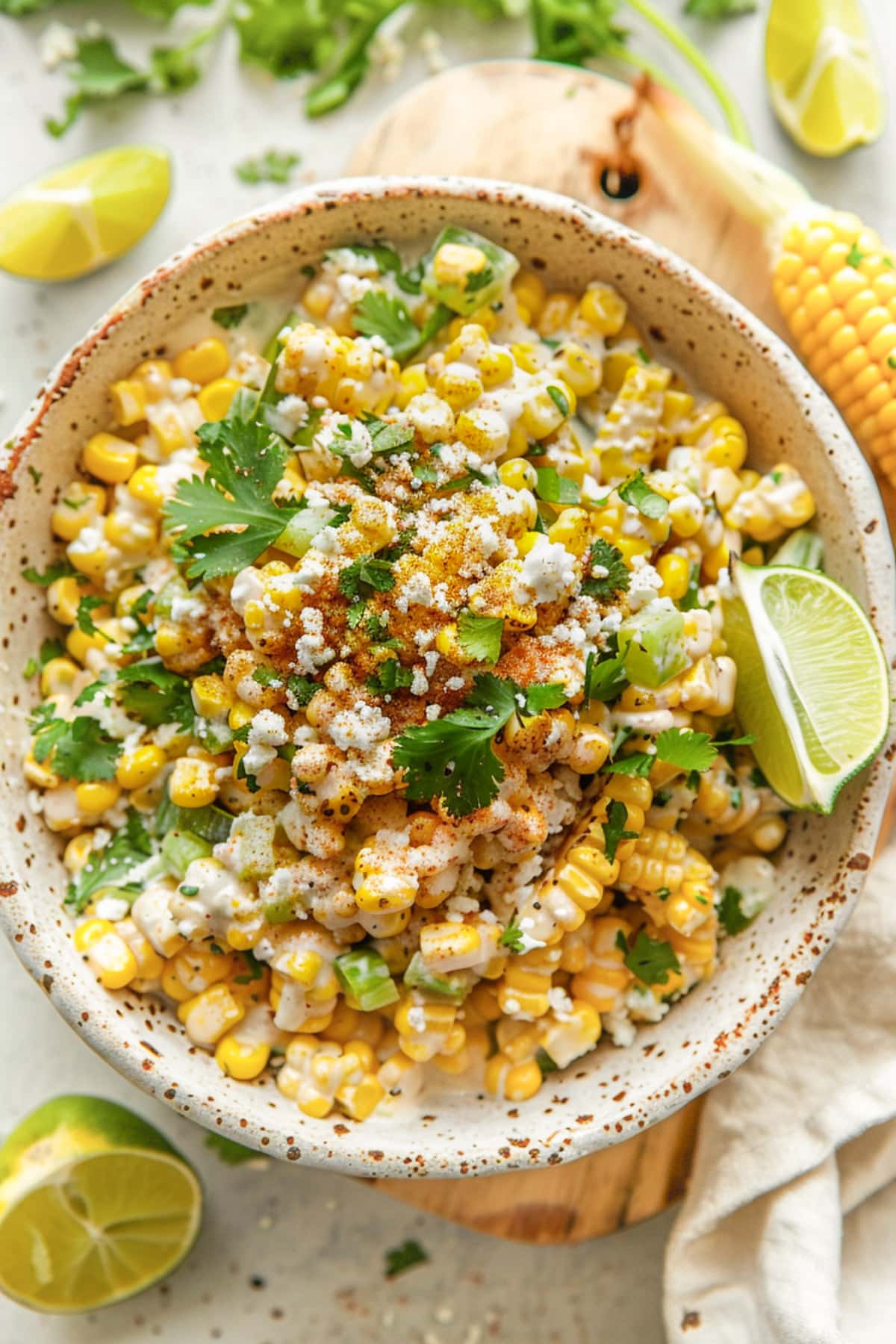 Bowl of Mexican street corn salad topped with crumbled cheese, chopped cilantro, green onions, and a sprinkle of chili powder, served with lime wedges on the side.