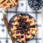 Whole Wheat Blueberry Oat Waffles on a blue plaid tablecloth with blueberries scattered around.
