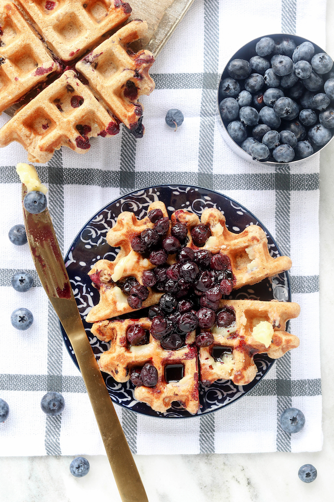 Whole Wheat Blueberry Oat Waffles on a blue plaid tablecloth with blueberries scattered around.