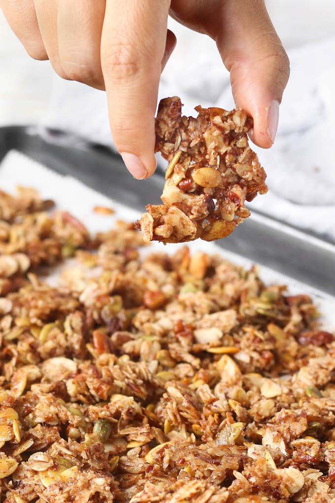 Heart and Seoul Grain Bowl Pumpkin Granola on a sheet pan. A hand is holding a large chunk of granola.