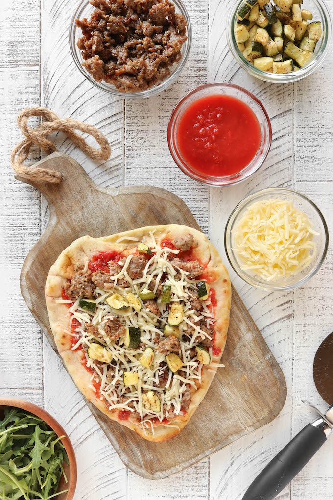 Chicken Sausage Flatbread Pizza on a wooden cutting board surrounded by pizza toppings.