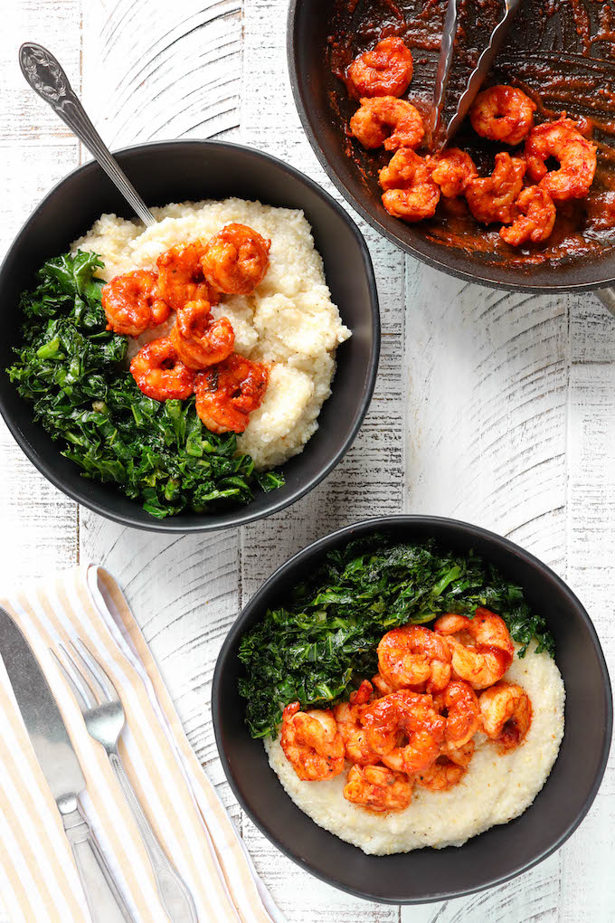 Cheddar BBQ Shrimp and Grits with Garlicky Greens in 2 black bowls sitting on a white surface.
