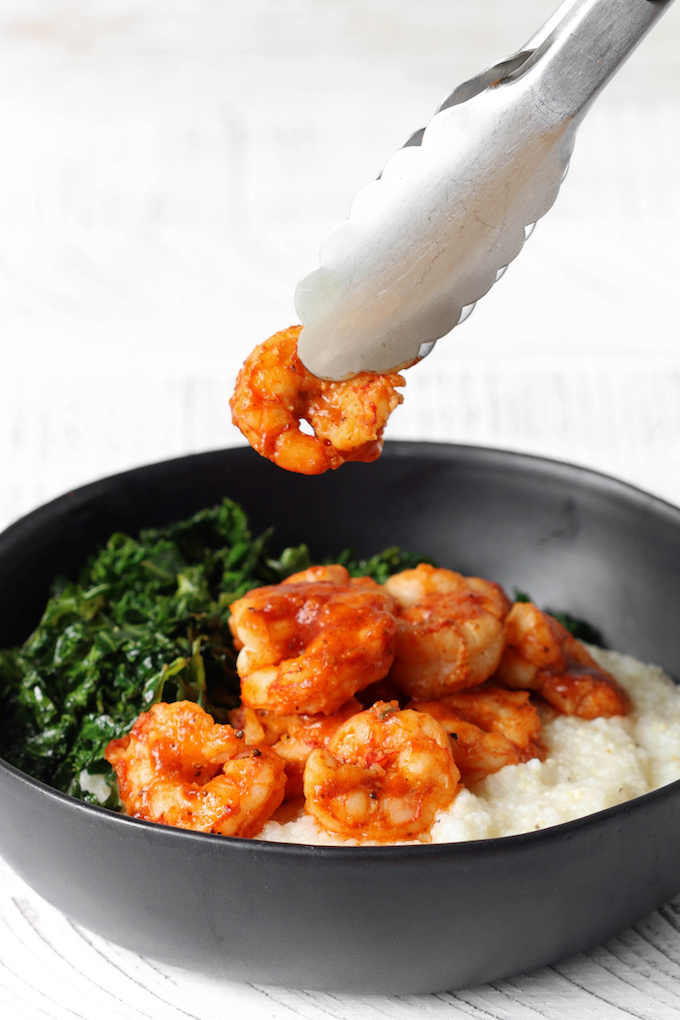 Cheddar BBQ Shrimp and Grits with Garlicky Greens in a black bowls. Shrimp piece is being lifted from bowl with silver and black tongs.