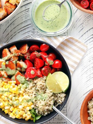 Chicken Burrito Bowl with Cilantro-Lime Dressing in a black bowl on a white wooden surface. A glass bowl of cilantro-lime dressing with a spoon in it is in the upper right hand corner.