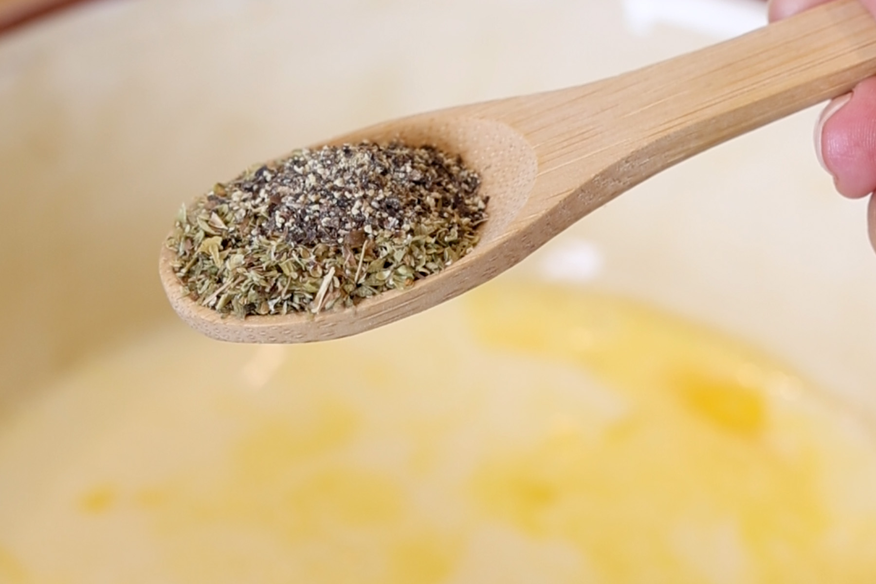 Wooden spoon holding oregano and black pepper over a bowl of beaten eggs for baked egg muffins.