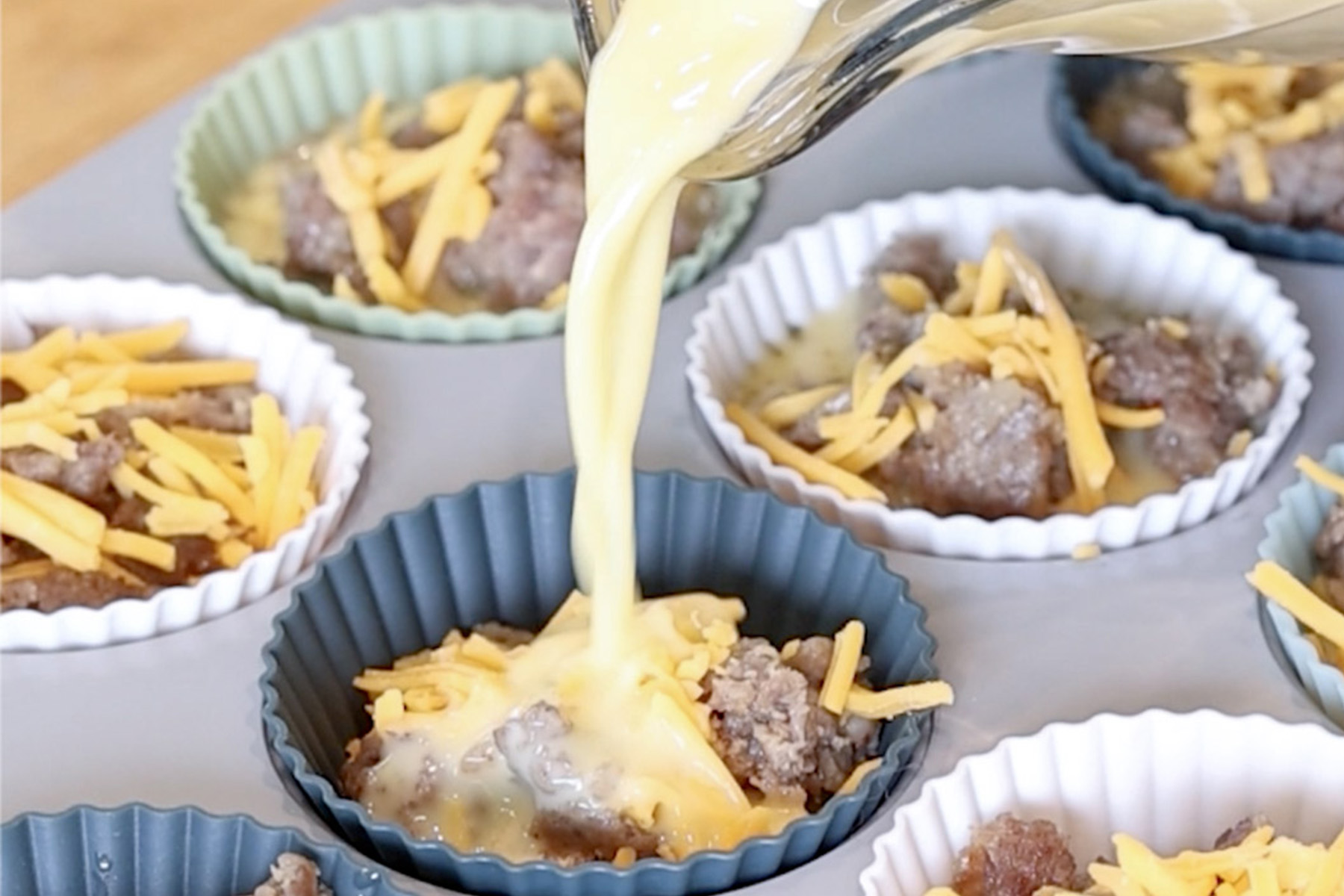 Egg mixture being poured over sausage and shredded cheddar in silicone muffin cups.