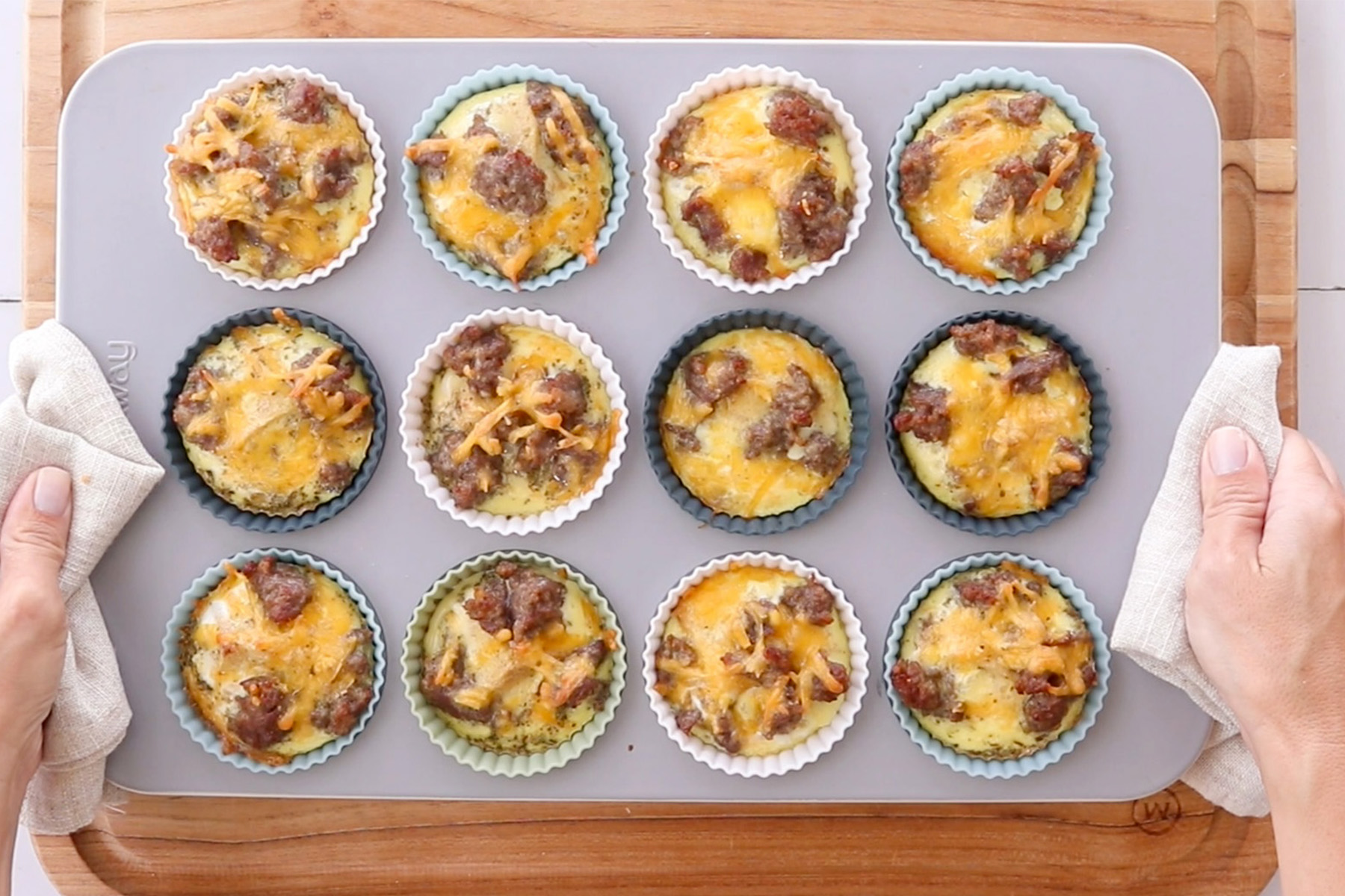Hands holding a tray of sausage and cheese egg muffins baked in colorful silicone cups.