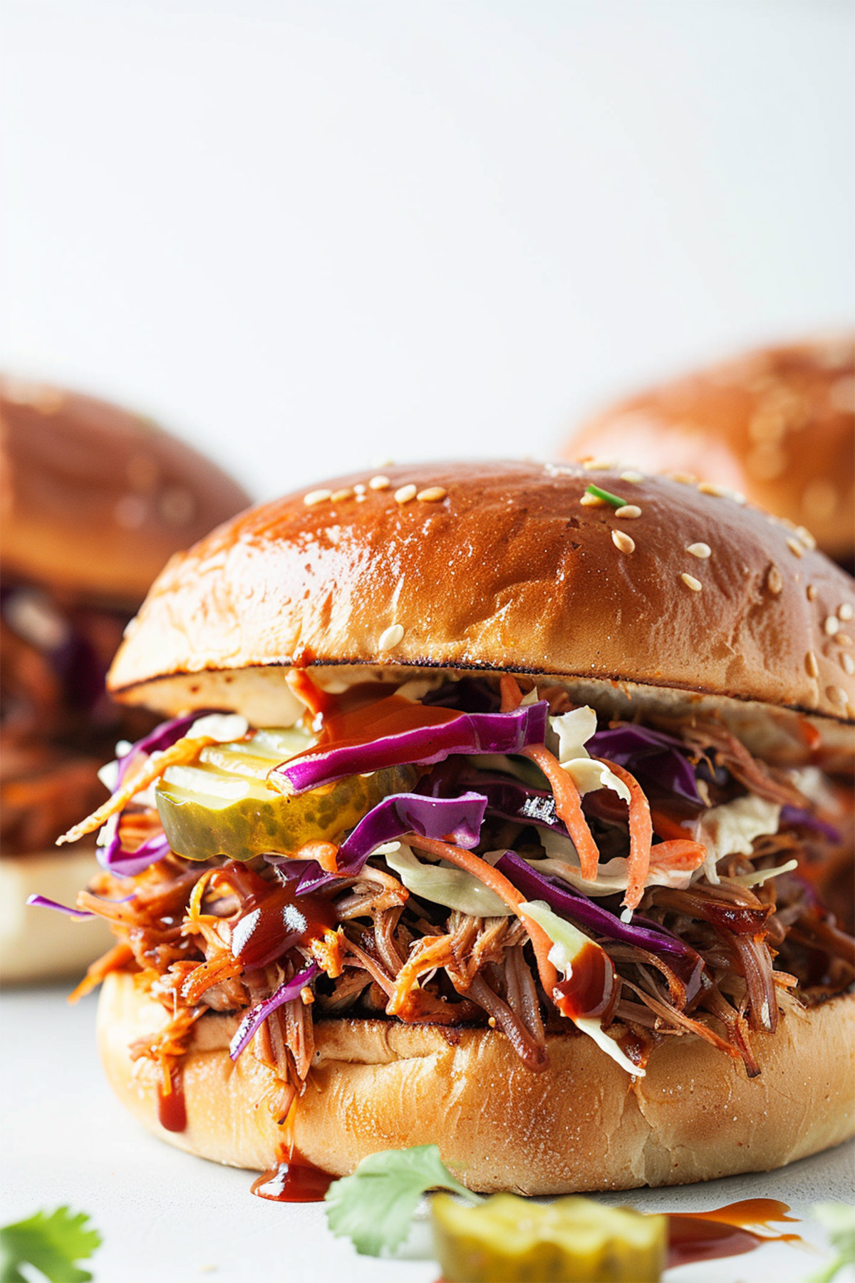 Close-up of a pulled pork sandwich on a toasted brioche bun, topped with barbecue sauce, crunchy coleslaw, and pickle slices, set against a bright white background.