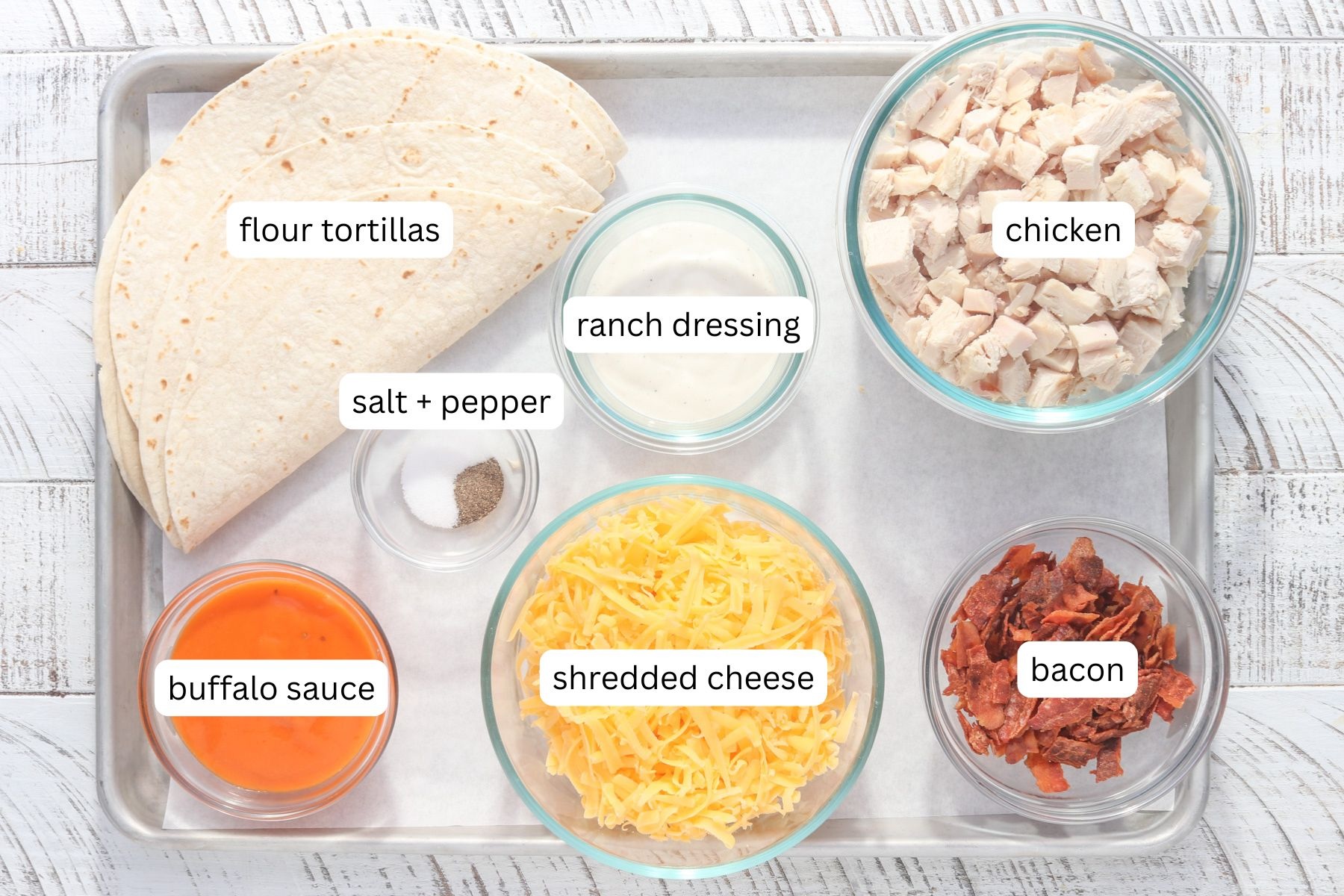 Overhead view of all ingredients needed for bacon chicken ranch quesadillas arranged on a sheet pan, including flour tortillas, diced cooked chicken, shredded cheddar cheese, crispy cooked bacon pieces, buffalo sauce, ranch dressing, and small bowls of salt and pepper.