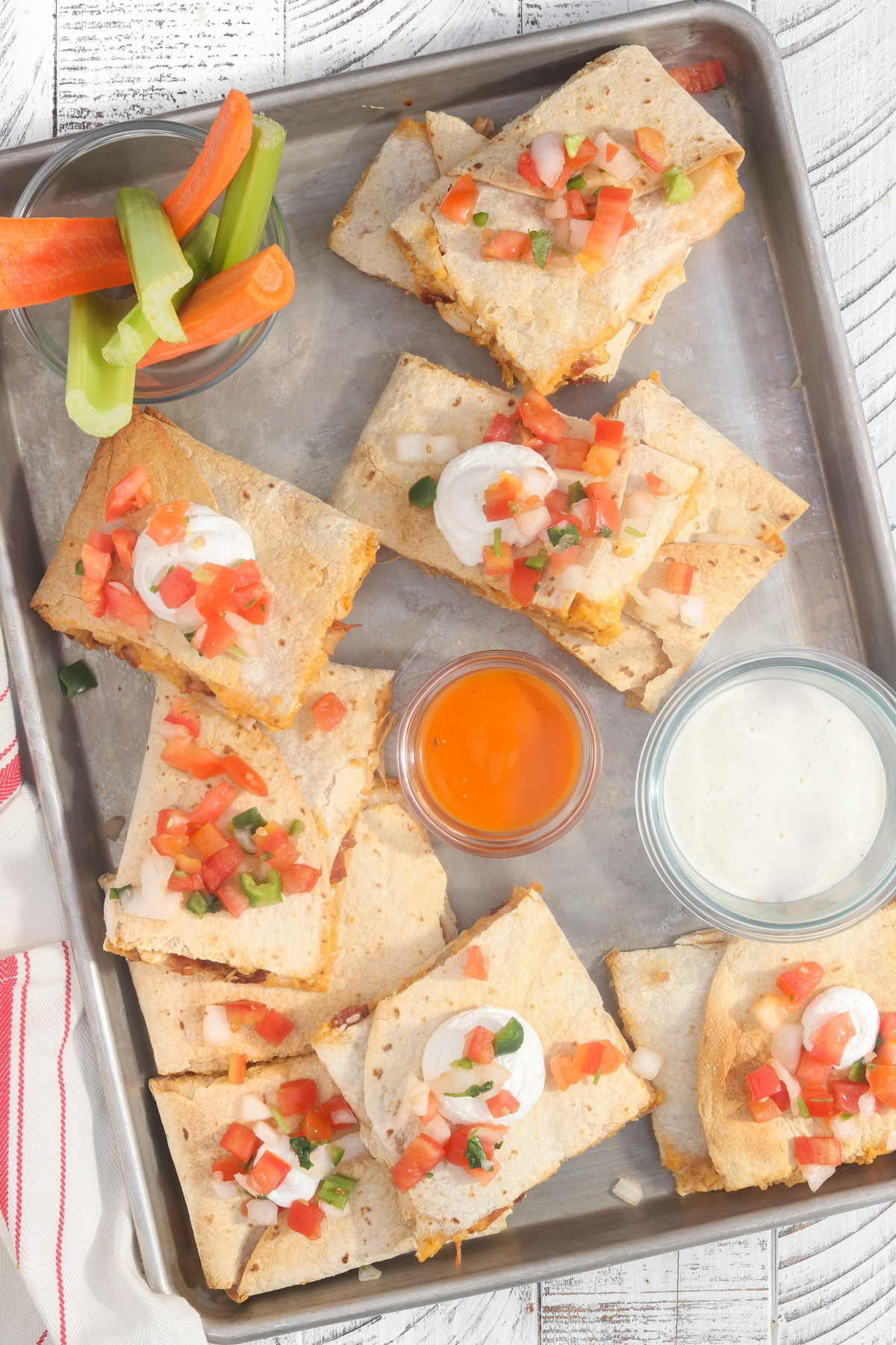 Tray of baked quesadilla slices topped with pico de gallo and sour cream, served with small bowls of buffalo sauce and ranch dressing, and a cup of celery and carrot sticks.