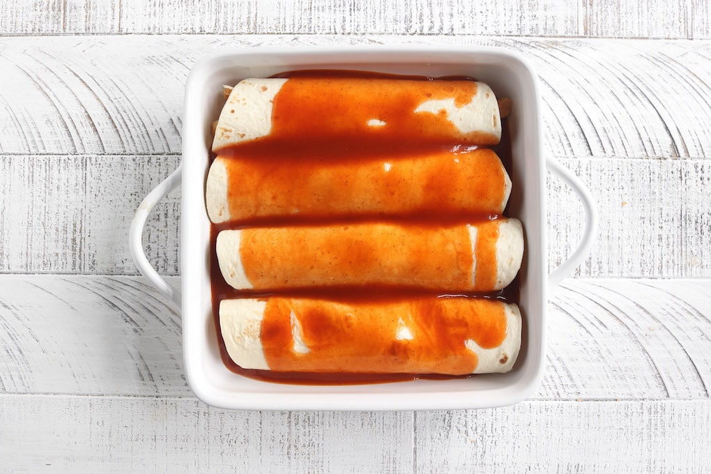 Step 5: A white baking dish filled with rolled chicken enchiladas topped with red enchilada sauce.