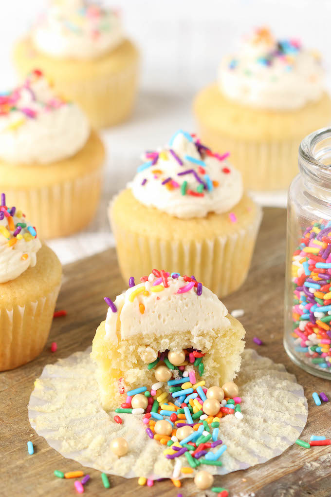 Sprinkle filled piñata cupcake with vanilla buttercream frosting sliced in half on a wood surface. Sprinkles are spilling from the center of the cupcake.