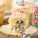 Sprinkle filled piñata cupcake with vanilla buttercream frosting sliced in half on a wood surface. Sprinkles are spilling from the center of the cupcake.