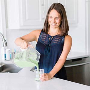 Bailey standing in a white kitchen pouring a smoothie out of a blender and into a glass.