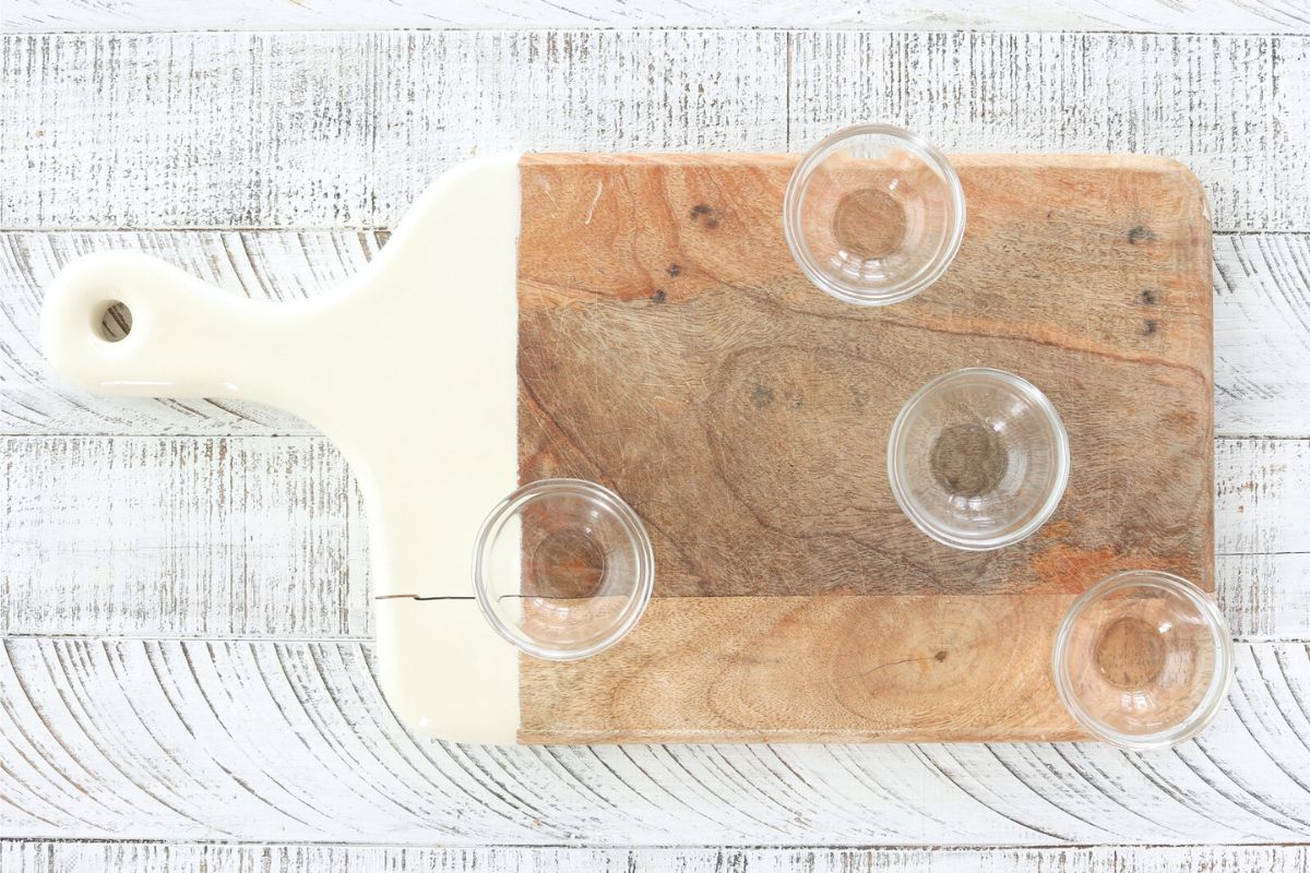 Step 1: Easy Charcuterie board. Wood cutting board with white handle on white background. 4 glass bowls arranged on board.