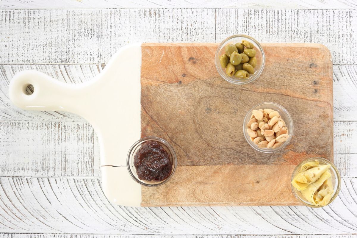 Cutting board with 4 bowls. Bowls are filled with olives, almonds, fig butter and artichokes.
